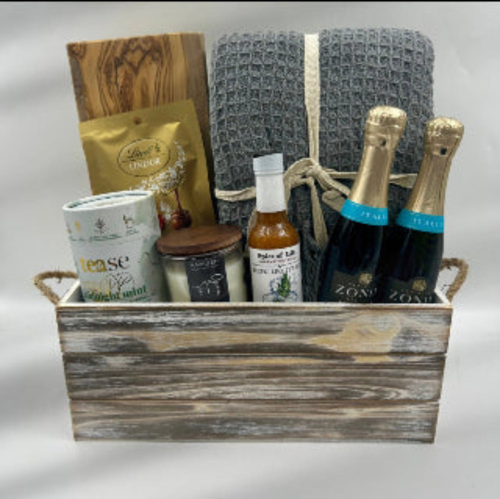 A cozy collection designed for moments of pure relaxation. This beautifully rustic wooden container holds an acacia cutting board for elegant snacking, a soft blanket for unwinding, and a lightly scented all-natural candle to set the mood. Sip premium tea or toast to special moments with two bottles of sparkling wine, while hot sauce and gourmet truffles add a dash of spice and sweetness to the experience.  Celebration Gift Baskets in Ottawa